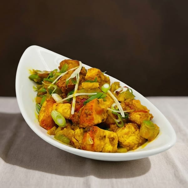 A white boat-shaped dish filled with golden paneer, green peppers, and scallions on a white tablecloth.