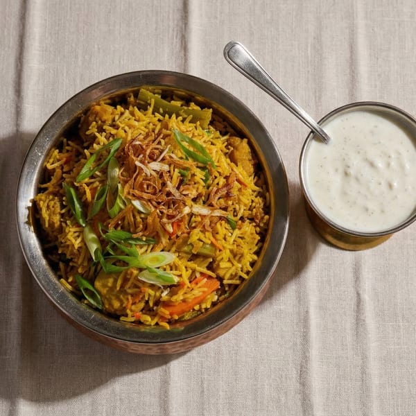 A bowl of chicken biryani with fried onions and green onions, served with raita.
