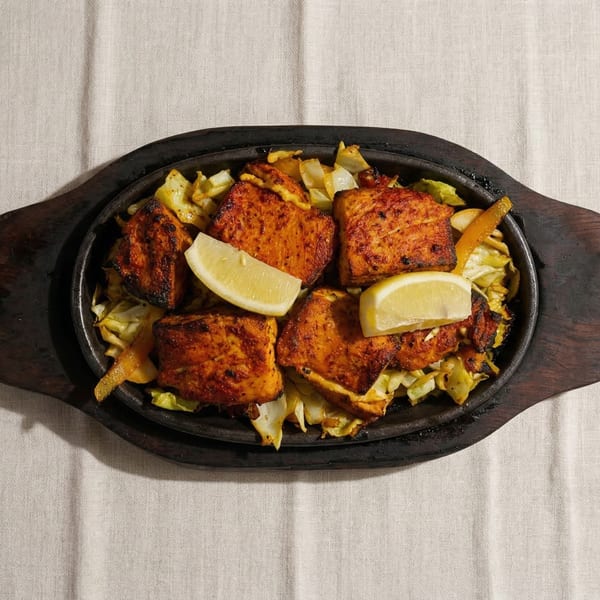 Sizzling salmon tikka with cabbage and lemon wedges on a dark wooden platter.