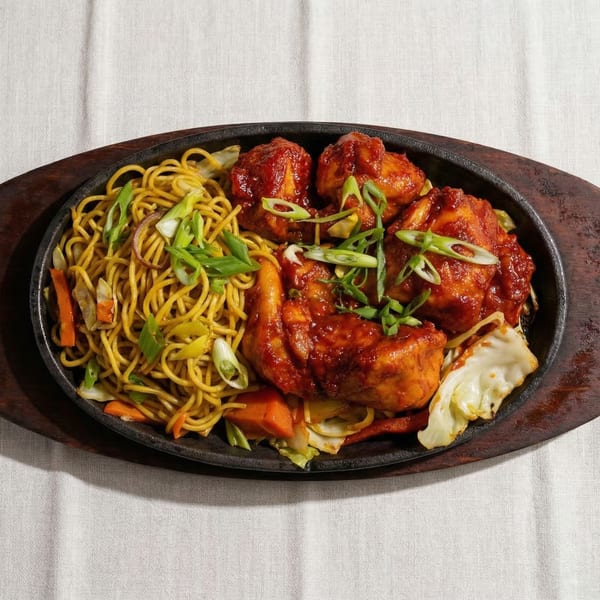 Sizzling platter with noodles, vegetables, and chicken in a rich red sauce, garnished with scallions.