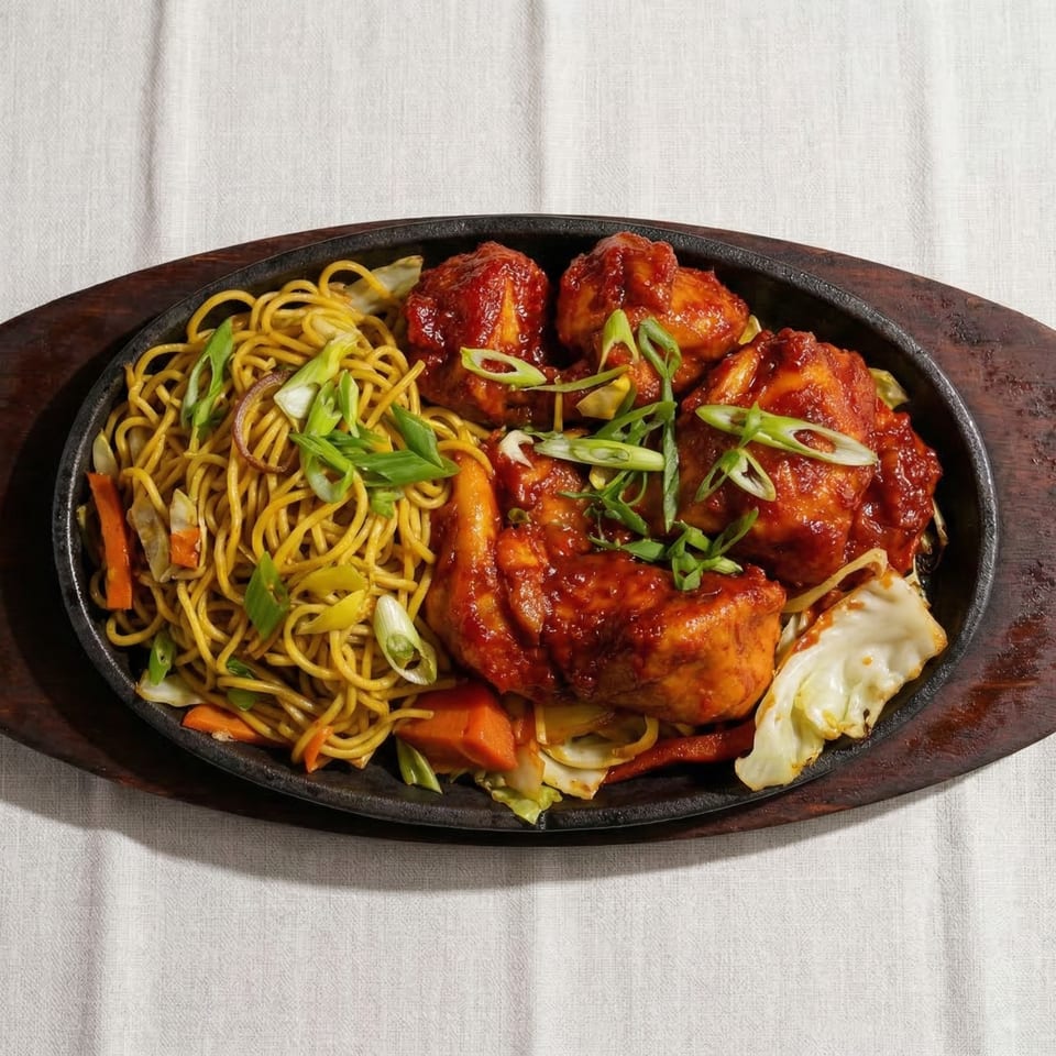 Sizzling platter with noodles, vegetables, and chicken in a rich red sauce, garnished with scallions.